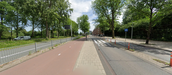 A very wide street consisting from right to left, a 2 lane car road (single direction), a grass raised divider, a 2 lane car road the other way, a fence, a 2 lane bidirectional bike lane, a side walk, a two lane bidirectional road and another sidewalk